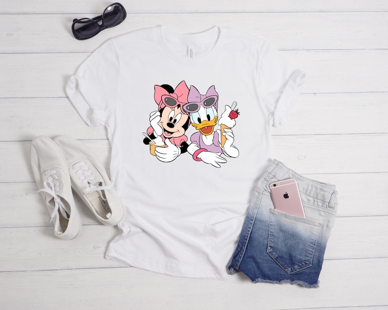 May include: A white short-sleeved t-shirt featuring a graphic print of Minnie Mouse and Daisy Duck wearing sunglasses and bows. Minnie holds a yellow cup with a spoon, while Daisy holds an ice cream cone topped with a strawberry.