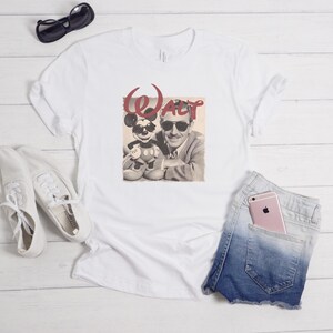 Puede incluir: Una camiseta blanca con un estampado gr&aacute;fico de Walt Disney y Mickey Mouse, ambos con gafas de sol, y el texto rojo "WALT" encima. La camiseta se muestra junto a unas zapatillas blancas, gafas de sol negras y unos pantalones cortos de mezclilla azules.