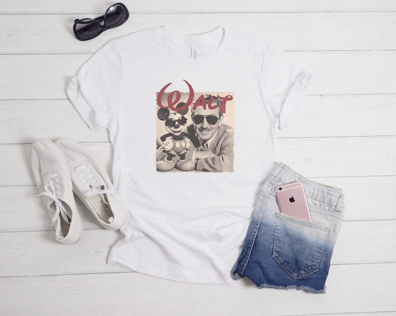 Puede incluir: Una camiseta blanca con un estampado gr&aacute;fico de Walt Disney y Mickey Mouse, ambos con gafas de sol, y el texto rojo "WALT" encima. La camiseta se muestra junto a unas zapatillas blancas, gafas de sol negras y unos pantalones cortos de mezclilla azules.
