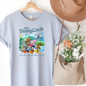 Op de afbeelding: Een lichtblauw t-shirt met een kleurrijke afbeelding van Walt Disney World bezienswaardigheden en personages, waaronder Mickey Mouse, Minnie Mouse, Donald Duck en Pluto. De tekst luidt Walt Disney World en The Most Magical Place on Earth.
