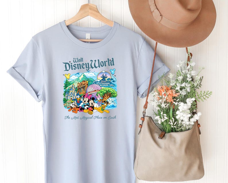 Op de afbeelding: Een lichtblauw t-shirt met een kleurrijke afbeelding van Walt Disney World bezienswaardigheden en personages, waaronder Mickey Mouse, Minnie Mouse, Donald Duck en Pluto. De tekst luidt Walt Disney World en The Most Magical Place on Earth.