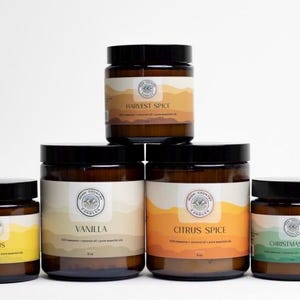 May include: Five amber glass candle jars with black lids. The candles are labelled with the scents: Citrus, Vanilla, Citrus Spice, Harvest Spice, and Christmas Tree.