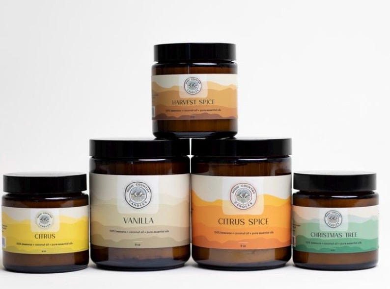 May include: Five amber glass candle jars with black lids. The candles are labelled with the scents: Citrus, Vanilla, Citrus Spice, Harvest Spice, and Christmas Tree.