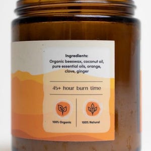 May include: A brown glass jar candle with a white label. The label reads "Ingredients: Organic beeswax, coconut oil, pure essential oils, orange, clove, ginger" and "45+ hour burn time". The label also features two icons, one stating "100% Organic" and the other "100% Natural".