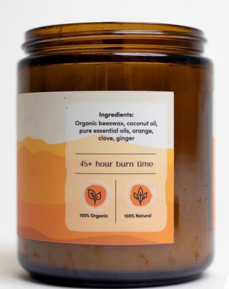 May include: A brown glass jar candle with a white label. The label reads "Ingredients: Organic beeswax, coconut oil, pure essential oils, orange, clove, ginger" and "45+ hour burn time". The label also features two icons, one stating "100% Organic" and the other "100% Natural".