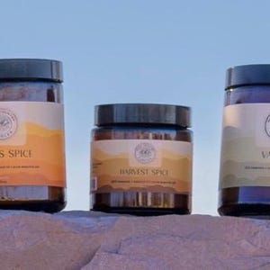 May include: Five glass jars with brown lids and labels. The labels are white with brown text and a logo. The jars are arranged in a row on a brown rock surface. The labels read "Citrus", "Citrus Spice", "Harvest Spice", "Vanilla", and "Cinnamon Hot".