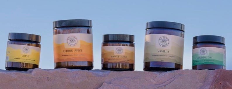 May include: Five glass jars with brown lids and labels. The labels are white with brown text and a logo. The jars are arranged in a row on a brown rock surface. The labels read "Citrus", "Citrus Spice", "Harvest Spice", "Vanilla", and "Cinnamon Hot".