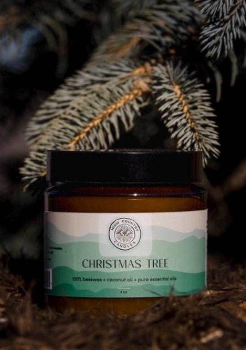 May include: A brown glass jar candle with a white label that says "High Country Candles" and "Christmas Tree". The label also says "100% beeswax + coconut oil + pure essential oils". The candle is sitting on a bed of pine needles.