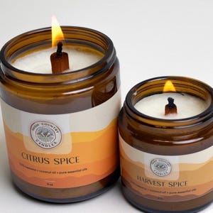 May include: Two amber glass candle jars with wooden wicks. The larger jar is labelled "Citrus Spice" and the smaller jar is labelled "Harvest Spice". Both jars are filled with white wax and have a label with a mountain and tree design.