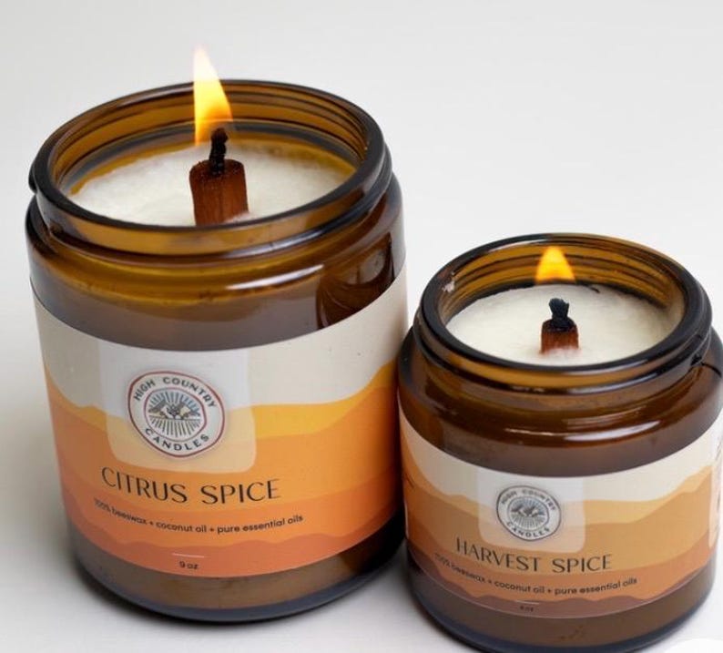 May include: Two amber glass candle jars with wooden wicks. The larger jar is labelled "Citrus Spice" and the smaller jar is labelled "Harvest Spice". Both jars are filled with white wax and have a label with a mountain and tree design.