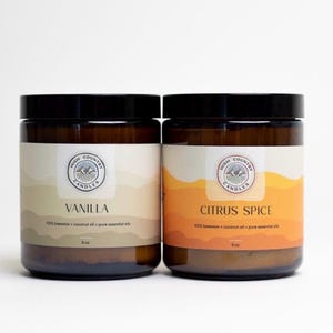May include: Two amber glass candle jars with black lids. The labels are white with brown text and a logo that says "High Country Candles". The left jar is labelled "Vanilla" and the right jar is labelled "Citrus Spice". Both jars are 9 oz.