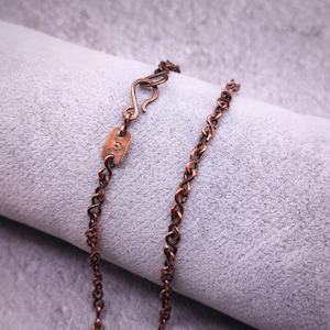 Puede incluir: Un collar de cadena de cobre con una pequeña etiqueta de cobre que tiene la letra "S" grabada.