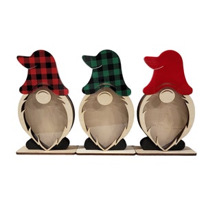 May include: Three wooden gnome figurines with different colored plaid hats. The gnomes are standing on a wooden base and have a cutout for a face.