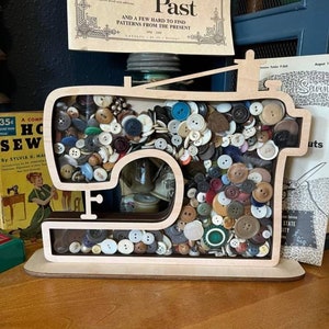 May include: A wooden sewing machine shaped box filled with a variety of colorful buttons. The box is sitting on a wooden surface. The box is a decorative storage container for buttons.