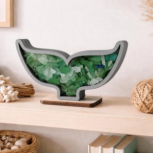 May include: A whale tail-shaped display frame with a gray border, filled with a collection of smooth, translucent sea glass pieces in various shades of green, white, and blue, mounted on a flat, rectangular wooden base for stability.