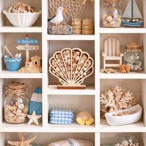 May include: A white display shelf filled with beach-themed decor. Items include seashells, a miniature sailboat, a seagull figurine, a wooden sign, a small beach chair, and a decorative wooden shell. The color palette is neutral with blue and beige accents.
