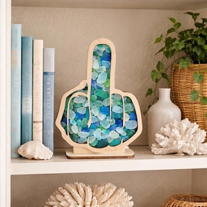 May include: A wooden sculpture in the shape of a hand giving the middle finger, filled with blue and green sea glass. The sculpture sits on a wooden base and is displayed on a white shelf with books and decorative items.