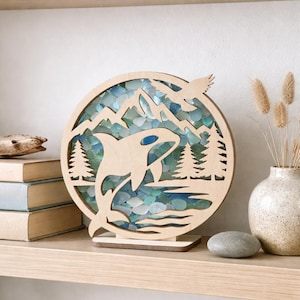May include: A wooden, circular art piece featuring a cutout design of an orca breaching the water, mountains, and trees. The background is filled with blue and green glass pieces. An eagle is cut out at the top. The piece sits on a wooden base.