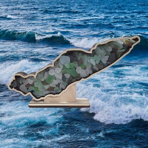 May include: A wooden cutout of an island filled with sea glass. The island is displayed on a wooden base against a blue ocean background.
