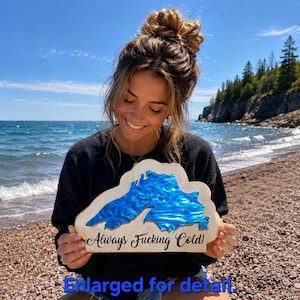 May include: A wooden cutout shaped like Lake Superior with a vibrant blue resin inlay representing the water. The light-colored wood features the black cursive text Always Fucking Cold! printed across the bottom section of the map shape.