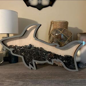May include: A wooden shark-shaped shadow box with a gray border, filled with dark shark teeth. The shark is displayed on a wooden surface, with a lamp and decorative items in the background. The word "Relax" is on a small wooden house.