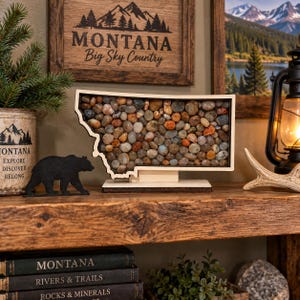 May include: A wooden display piece shaped like the state of Montana, filled with a collection of smooth, multi-colored river stones. The object is mounted on a light-colored wooden base and features a dark frame outlining the state's border.