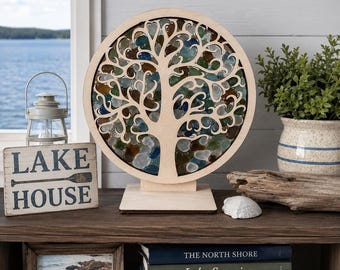 May include: A circular light-colored wooden frame featuring a laser-cut tree silhouette filled with translucent blue, green, and brown sea glass pieces. The tree is mounted on a rectangular wooden base and stands upright as a decorative display piece.