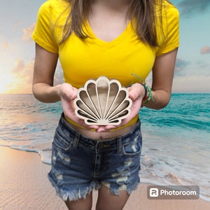 May include: A wooden seashell shaped box with a cutout design. The box is held in the hands of a person wearing a yellow shirt and denim shorts.