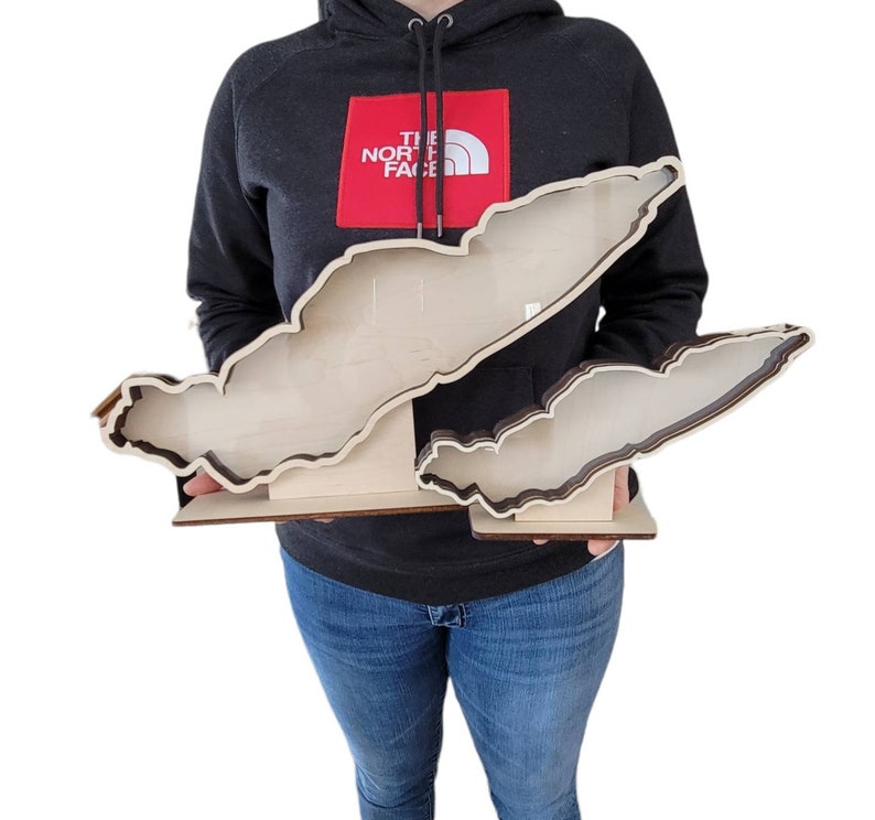 May include: A wooden cutout of the Great Lakes, with each lake individually cut out and layered. The cutout is held by a person wearing a black jacket with a red "The North Face" logo.