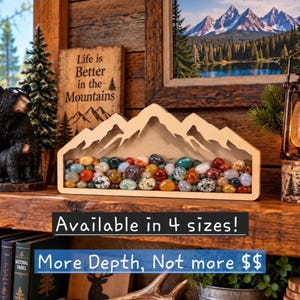May include: A light-colored wooden mountain-shaped display frame filled with a collection of smooth, multi-colored polished stones. The frame features a cutout mountain silhouette at the top. Text on the image reads: Available in 4 sizes! More Depth, Not more $$.