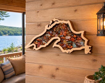 May include: A wooden wall art piece shaped like the outline of Lake Superior, filled with a collection of polished, multi-colored stones in shades of red, orange, brown, and grey, mounted on a horizontal wood-paneled wall surface.