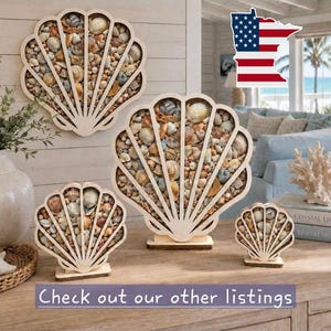 May include: Three light-colored wooden shell-shaped display frames of varying sizes, each filled with a collection of assorted natural seashells in shades of tan, brown, white, and grey, standing upright on flat rectangular wooden bases.