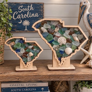 May include: Two wooden South Carolina state-shaped display frames filled with multicolored sea glass and small seashells. Each frame is mounted on a rectangular wooden base, showcasing a collection of blue, green, brown, and clear glass pieces.