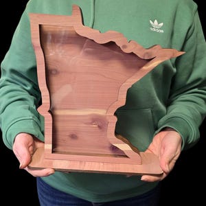 May include: A wooden cutout of the state of Minnesota, with a clear acrylic insert, is being held by a person wearing a green hoodie with the Adidas logo. The cutout is made of cedar wood and has a natural wood finish.