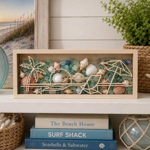 May include: A rectangular wooden shadow box frame containing a layered beach scene. The display features a laser-cut wooden palm tree, a sun, a beach chair, and an umbrella, all set against a bed of small, multi-colored sea glass and seashells.