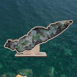 May include: A wooden cutout of an island filled with sea glass. The island is on a wooden stand. The background is a teal blue.