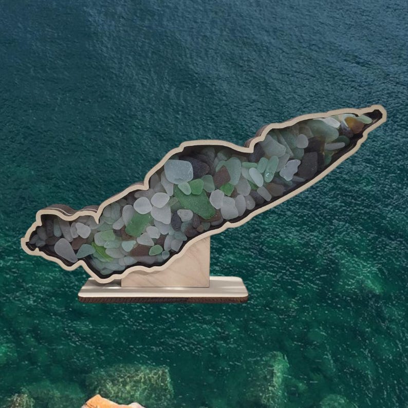 May include: A wooden cutout of an island filled with sea glass. The island is on a wooden stand. The background is a teal blue.