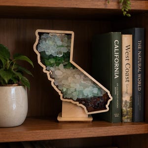 May include: A wooden display frame shaped like the state of California, filled with layers of smooth sea glass in shades of clear, white, light green, dark green, and dark brown, supported by a small wooden base stand on a shelf.