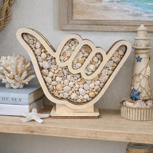 May include: Wooden decorative 'Shaka' hand sign filled with various seashells. The sign is displayed on a wooden shelf with other coastal decor, including a small lighthouse and a framed beach scene painting.