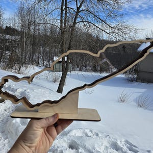 May include: A wooden cutout of a lake in the shape of a state, standing on a wooden base. The cutout is brown and the base is a light wood color. The cutout is in front of a snowy background.