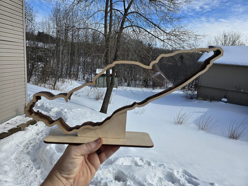May include: A wooden cutout of a lake in the shape of a state, standing on a wooden base. The cutout is brown and the base is a light wood color. The cutout is in front of a snowy background.