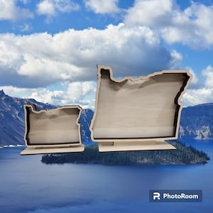 May include: Two wooden cutout shapes of the state of Oregon, one larger than the other, both standing on a wooden base. The cutouts are light brown and have a natural wood finish. The background is a blue sky with white clouds.