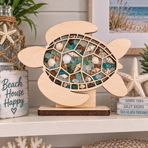 May include: A light-colored wooden turtle-shaped display frame featuring a hollow shell section filled with various small seashells and smooth blue, green, and brown sea glass pieces, supported by a rectangular wooden base stand.