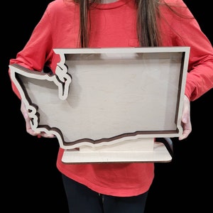 May include: A wooden cutout of the state of Washington with a clear acrylic insert. The cutout is on a wooden stand and is being held by a person.