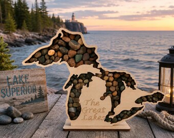 May include: A light-colored wooden display case shaped like the Great Lakes, filled with various small, smooth, multi-colored stones. The wood features the engraved text 'The Great Lakes' on the front surface of the display.