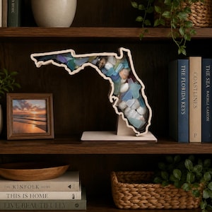 May include: A wooden display piece shaped like the state of Florida, featuring a clear front panel filled with various sea glass pieces and small white seashells. The object is mounted on a flat wooden base and stands upright on a shelf.