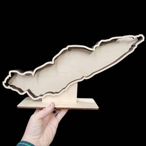 May include: A wooden cutout of an island with a brown outline and a natural wood finish. The cutout is mounted on a small wooden base.
