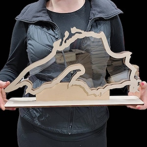 May include: A layered wooden sculpture of a geographical region, possibly a lake or coastline, with a clear acrylic insert. The sculpture is mounted on a rectangular wooden base. The wood has a natural, light brown color.