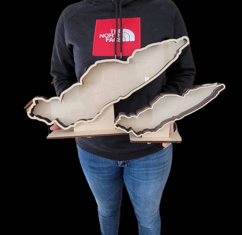 May include: A wooden cutout of Lake Champlain, a large lake on the border of New York, Vermont, and Quebec, Canada. The cutout is made of light wood with a dark brown outline. The cutout is held by a person wearing a black sweatshirt with a red "The North Face" logo.