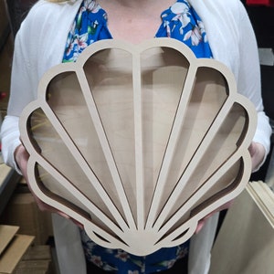 Puede incluir: Una caja de exhibici&oacute;n de madera clara con forma de concha marina. La parte frontal presenta un panel transparente enmarcado por listones de madera que irradian desde la base, creando secciones distintas dentro de la silueta de la concha.